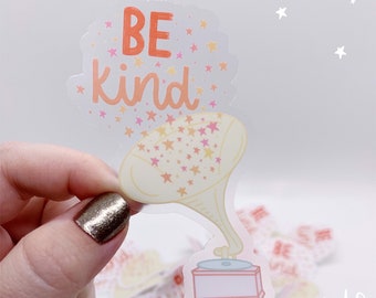 Puede incluir: Una pegatina transparente con un diseño de tocadiscos vintage. El tocadiscos está reproduciendo un disco con las palabras "Be Kind" en letras rosa y naranja. La pegatina está rodeada de pequeñas estrellas rosas y amarillas.