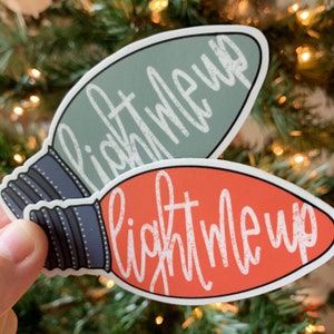 May include: Two stickers in the shape of Christmas light bulbs. One is orange with white text that reads "Light Me Up." The other is a muted green with the same text. The bulbs have a black and gray base.