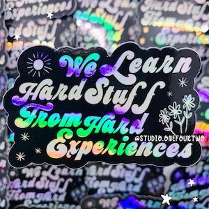 May include: A black and iridescent holographic sticker with the text "We Learn Hard Stuff From Hard Experiences" in a white, retro font. The sticker also features a small sun, two white flowers, and the text "@STUDIO.ONEFOURTWO" in a smaller font.