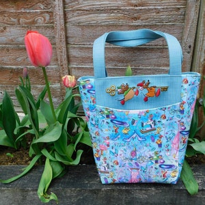 May include: A light blue tote bag with a whimsical design featuring a Cinderella theme. The bag has a large pocket on the front with an embroidered image of three mice holding a key.