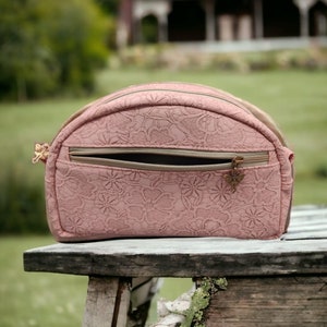 Puede incluir: Un bolso de mano de cúpula rosa con estampado floral, cremallera dorada y un pequeño charm de mariposa de bronce decorativo.