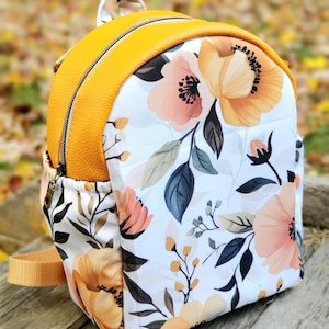 May include: A small yellow and white floral backpack with a zip closure. The backpack has a white background with a floral pattern in shades of pink, yellow, and orange. The top of the backpack is made of yellow faux leather.