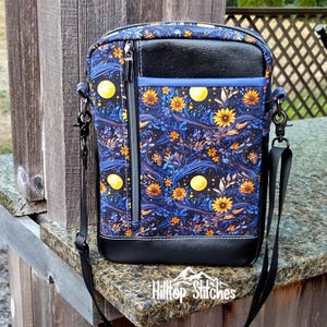 May include: A blue and black crossbody bag with a floral pattern and yellow moons. The bag has a zipped pocket and a black leather flap. The bag is made by Hilltop Stitches.