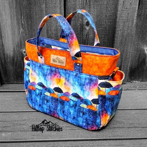 May include: A handmade tote bag with a vibrant design. The bag features a blue fabric with a pattern of umbrellas in various colors. The top section is orange, and the handles are patterned with a galaxy design. The text "Hilltop Stitches" is visible on the bag.
