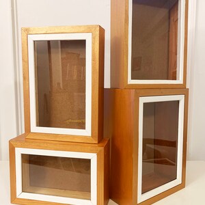 May include: Four wooden shadow boxes with white trim. The boxes are stacked on top of each other, with the largest box on the bottom and the smallest box on top. The boxes are all made of a light brown wood and have a clear glass front.