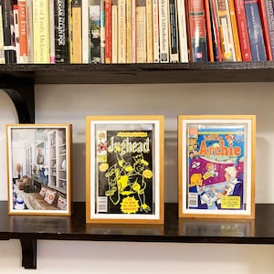May include: Three framed pictures on a shelf. The picture on the left shows a room with a couch and a bookshelf. The middle picture shows a comic book cover featuring Jughead. The picture on the right shows a comic book cover featuring Archie.