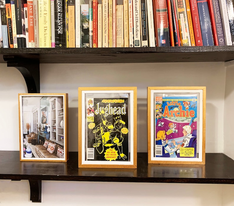 May include: Three framed pictures on a shelf. The picture on the left shows a room with a couch and a bookshelf. The middle picture shows a comic book cover featuring Jughead. The picture on the right shows a comic book cover featuring Archie.