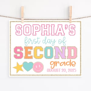 以下が含まれることがあります： ピンク、青、オレンジ、紫で「SOPHIA'S first day of SECOND grade」と書かれたカラフルなサイン。星、ハート、スマイリーフェイスのグラフィックと「AUGUST 20, 2025」の日付が含まれています。木製のクリップで吊るされています。