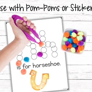 May include: A printable worksheet for learning the letter "U" with a picture of a horseshoe and the text "U is for horseshoe." The worksheet has a grid of circles for placing pom-poms or stickers. A hand is shown using tweezers to place a pink pom-pom on the worksheet. A bowl of colorful pom-poms is shown to the right of the worksheet.