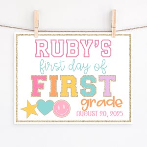 May include: A decorative sign with the text "RUBY'S first day of FIRST grade AUGUST 20, 2025" in pink, blue, orange, yellow, and purple. The sign is decorated with a gold glitter border, a star, a heart, and a smiley face.