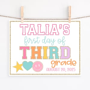 May include: A colorful sign with the text "TALIA'S first day of THIRD grade AUGUST 20, 2025". The sign has a gold glitter border and is decorated with a star, heart, and smiley face.