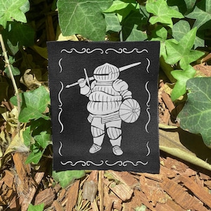 May include: A rectangular black fabric patch featuring a white screen-printed illustration of a knight in full plate armor. The knight holds a long sword across their shoulders and carries a round shield. A decorative white border frames the design.