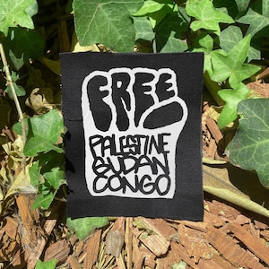 May include: A black square patch with the word "FREE" in bold white letters above a white fist. Below the fist, the words "PALESTINE SUDAN CONGO" are printed in white.