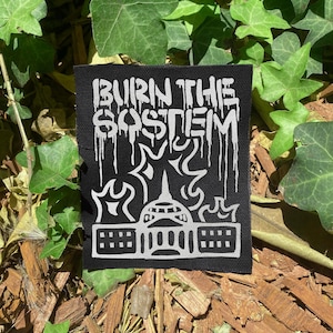 Op de afbeelding: Zwart-wit patch met de tekst "BURN THE $OSTEM" in een druipend lettertype. Het ontwerp bevat een gestileerd gebouw met vlammen, tegen een zwarte achtergrond. De patch is omgeven door groene bladeren en houtsnippers.