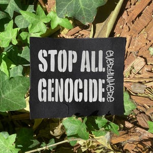 Könnte beinhalten: Schwarzes quadratisches Patch mit weißem Text, der "STOP ALL GENOCIDE" in fetten Blockbuchstaben lautet. Das Wort "everywhere" ist vertikal auf der rechten Seite aufgedruckt. Der Patch ist von grünen Blättern und Holzspänen umgeben.