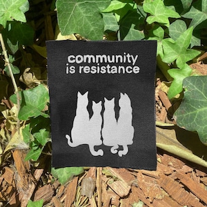 May include: A rectangular black fabric patch featuring the white text "community is resistance" printed above a graphic of four white cat silhouettes sitting side by side. The patch has a visible woven texture and clean, straight edges.