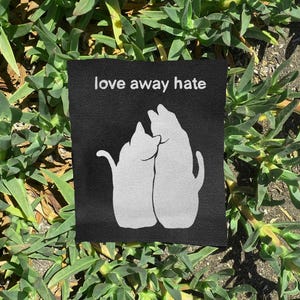 May include: A rectangular black fabric patch featuring a white graphic of two cats sitting side by side. Above the cats, the text "love away hate" is printed in a simple white sans-serif font. The patch has a visible woven texture throughout.