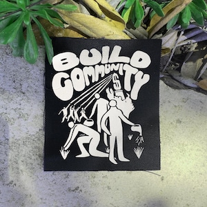 Puede incluir: Parche cuadrado negro con las palabras "BUILD COMMUNITY" en blanco, con letras estilizadas. El parche presenta un gráfico de varias figuras, algunas extendiéndose hacia rayos de luz y otras cuidando plantas.