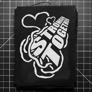 May include: Black square patch with the words "STRONGER TOGETHER" in white, stylized text. The design includes a heart and cloud-like shapes. The patch is on a black grid.