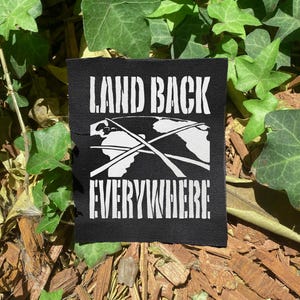 May include: A black patch with the white text "LAND BACK EVERYWHERE" and a graphic of the Earth with lines. The patch is set against a backdrop of green leaves and brown mulch.