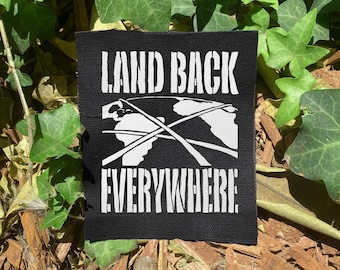 May include: Black square patch with white text reading "LAND BACK EVERYWHERE" above a graphic of the Earth with lines crossing it. The patch is surrounded by green leaves and brown mulch.