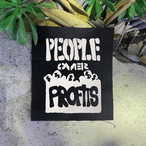May include: Black square patch with white text that reads "PEOPLE OVER PROFITS." The text is in a bold, stylized font. The word "PROFITS" is above a graphic of money bags.