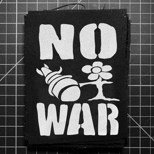 May include: Black fabric patch with white text and graphics. The words "NO WAR" are displayed prominently. A stylized bomb and flower are featured in the center. The patch is on a black grid background.