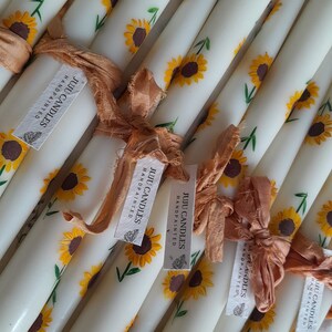 May include: A set of white taper candles hand-painted with yellow sunflowers. The candles are tied with brown ribbon and have the text "JUIJU CANDLES HANDPAINTED" on the labels.