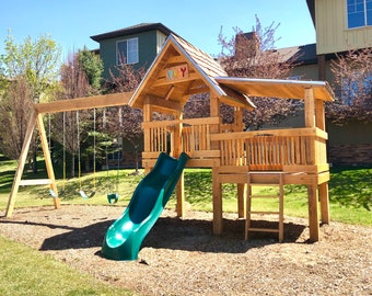May include: A wooden playground set with a slide, swing set, and a small wooden house with the word "PLAY" on the front. The set is in a grassy area with a gravel surface.