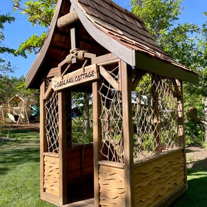 May include: A wooden playhouse with a shingled roof and the words "WOODLAND COTTAGE" on a sign. The structure features rope netting and a decorative wave pattern on the lower panels, set on a grassy lawn.