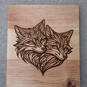 Peut inclure: Planche à découper en bois avec un motif pyrogravé de deux renards se faisant face. Les renards sont représentés avec des motifs et des lignes complexes, créant une image stylisée et artistique. Le bois est de couleur brun clair naturel.