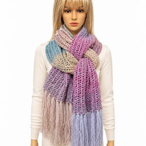 May include: A chunky knit scarf featuring a variegated color palette of purple, blue, beige, and pink. The scarf has a ribbed texture and long, straight fringe detailing at both ends, creating a layered look around the neck area.
