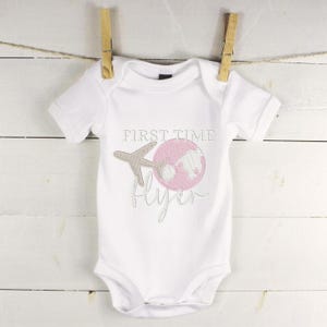 Könnte beinhalten: Weißer Baby-Strampler mit dem gestickten Text "First Time Flyer" in Braun und Rosa. Das Design beinhaltet einen rosafarbenen Globus mit einem braunen Flugzeug.