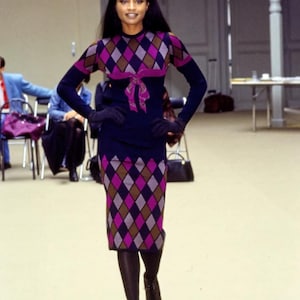 Könnte beinhalten: Eine Frau trägt ein schwarzes und violettes Kleid mit Rautenmuster und einer schwarzen Schleife in der Taille. Sie trägt schwarze Strumpfhosen und schwarze Stiefel mit Absatz.