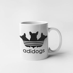 Puede incluir: Taza de cerámica blanca con una silueta negra de tres perros en formación triangular, que se asemeja al logotipo de Adidas. El texto "adidogs" está impreso debajo del logotipo.