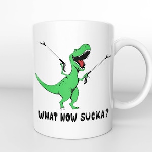 Op de afbeelding: Witte keramische mok met een groene dinosaurus die twee stokken vasthoudt en de tekst "WHAT NOW SUCKA?" gedrukt op de voorkant.