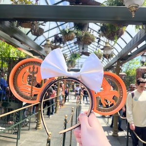 May include: Orange and gold Mickey Mouse ears with a white bow. One ear features a gold trident and waves, the other ear features a gold Pegasus.