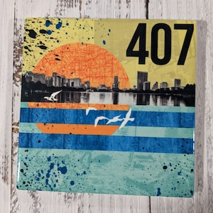 Puede incluir: Un azulejo cuadrado amarillo, azul y naranja con una silueta de ciudad y una puesta de sol. El azulejo tiene el número 407 impreso en él.