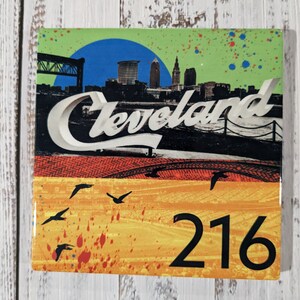 May include: A square ceramic tile with a colorful design featuring a cityscape with the word "Cleveland" in white lettering. The tile has a yellow, orange, and red background with black birds flying across the bottom. The number "216" is printed in black in the bottom right corner.
