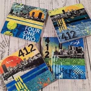 May include: Four ceramic coasters with a cityscape design. The coasters feature a yellow, blue, and green color scheme with a cityscape silhouette and the number 412. One coaster also has the words "Four One Two" on it.