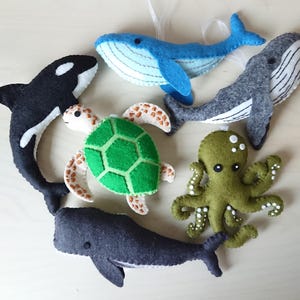 May include: A set of felt ocean animal ornaments, including a blue whale, a grey whale, a black and white orca, a green sea turtle, and a green octopus.