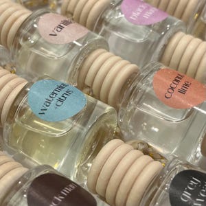 May include: A close-up of various car air fresheners with different scents. The air fresheners are in glass bottles with wooden caps and colorful labels. The labels have text that includes "Vanilla", "Peach Mango", "Watermint + Citrus", "Coconut Lime", and "Black Moon".