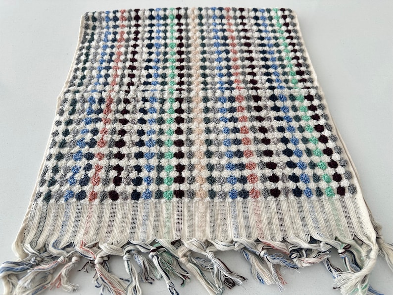 K&ouml;nnte beinhalten: Ein wei&szlig;es Handtuch mit einem bunten geometrischen Muster. Das Handtuch hat einen Fransenrand und ist aus einem weichen, saugf&auml;higen Material gefertigt.