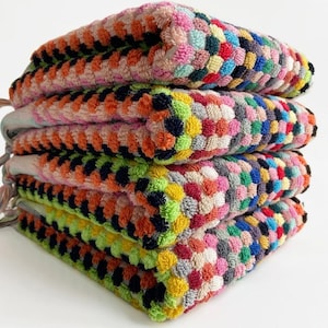 May include: A stack of colorful, square-shaped towels. Each towel features a textured, geometric pattern in a variety of colors, including pink, orange, green, blue, and yellow. The towels have small, decorative tassels on the side.
