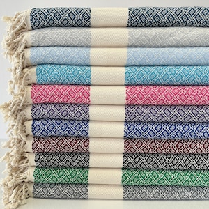 Op de afbeelding: Een stapel van tien Turkse handdoeken in verschillende kleuren, waaronder turquoise, roze, paars, blauw, bordeauxrood, zwart, groen en grijs. Elke handdoek heeft een witte streep in het midden en een ruitpatroon.