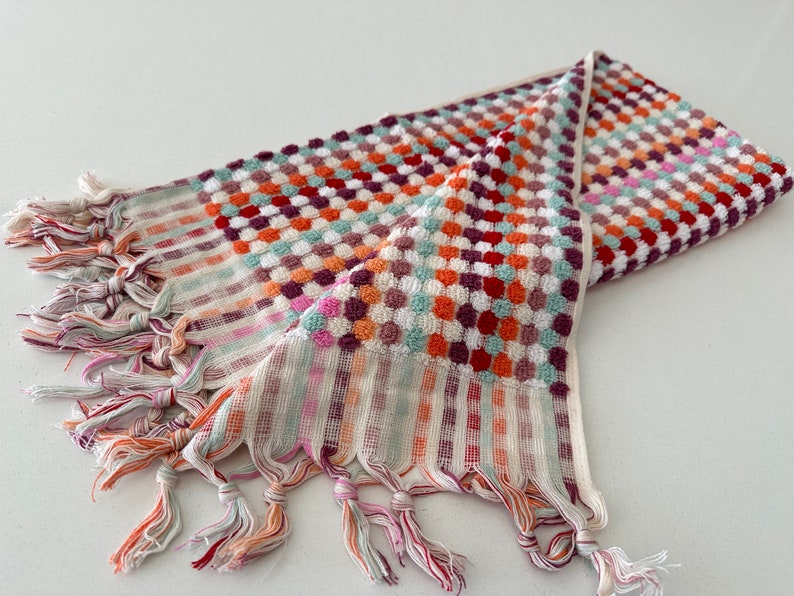 May include: A colorful hand towel with a checkered pattern and fringe. The towel is made of a soft, absorbent material and features a variety of colors, including red, orange, green, and purple.