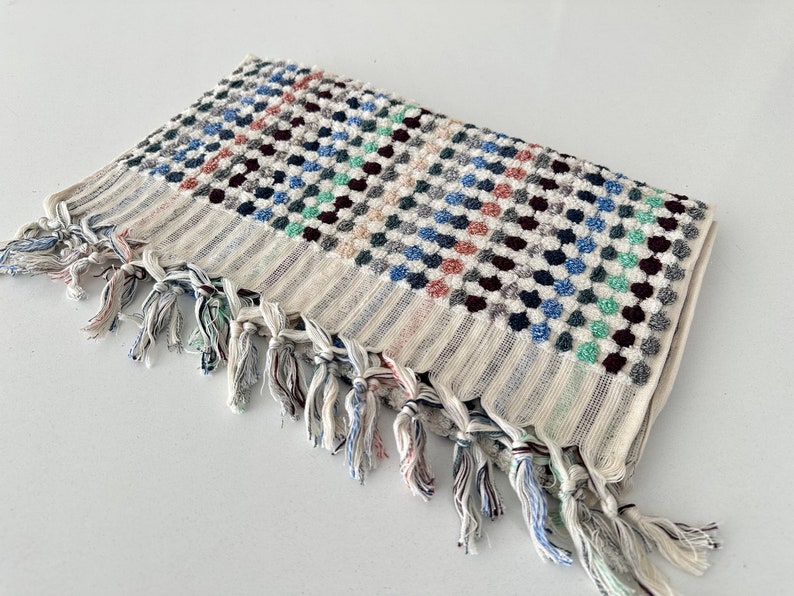 K&ouml;nnte beinhalten: Ein wei&szlig;es Badetuch mit einem bunten geometrischen Muster und Fransen. Das Muster besteht aus kleinen, erhabenen Kreisen in Blaut&ouml;nen, Gr&uuml;n, Rot und Schwarz.