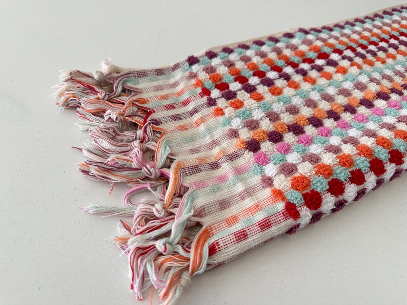 May include: A colorful striped and patterned bath towel with a fringe border. The towel is made of a soft, absorbent material and features a variety of colors, including red, orange, pink, green, and white.