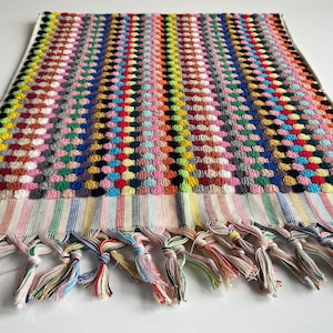 May include: A colorful, textured towel with a repeating pattern of raised dots in various colors. The towel has a striped border and knotted fringe in multiple colors. The towel is likely made of cotton and is suitable for use in a bathroom or at the beach.