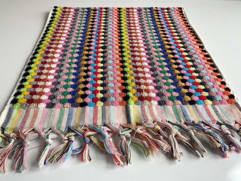May include: A colorful, textured towel with a repeating pattern of raised dots in various colors. The towel has a striped border and knotted fringe in multiple colors. The towel is likely made of cotton and is suitable for use in a bathroom or at the beach.
